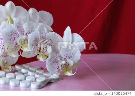Close-up of white pills in a blister against orchid flowers with lilac spots on a pink surface and red background. 143036474