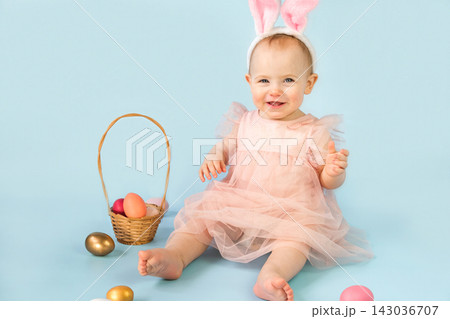 Happy baby girl, ten months old, wearing bunny ears headband, sitting on light blue studio background and playing with colored easter eggs. Little Child. Easter greeting card. Baby easter bunny 143036707