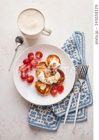Savory syrniki (cheese pancakes) topped with sour cream, crispy fried onions, and fresh cherry tomatoes, served on a white plate with a blue knit cloth. Perfect for breakfast or brunch. Savory syrniki (cheese pancakes) topped with sour cream, crispy fried onions, and fresh cherry tomatoes, served on a white plate with a blue knit cloth. Perfect for breakfast or brunch. 143038329