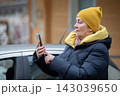 Woman wearing yellow hat checks smartphone outside her car in a city during winter 143039650