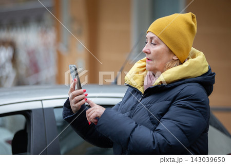 Woman wearing yellow hat checks smartphone outside her car in a city during winter 143039650