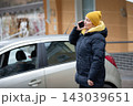 Woman in yellow hat talking on phone near parked car in urban setting during winter day 143039651