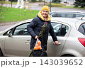 Woman carrying bag of oranges and holding umbrella while getting into car in urban area during daytime 143039652