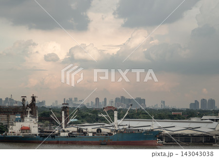Moored along the Chao Phraya River, a large industrial vessel sits framed by the lush foliage of Bang Kachao, distant city skyscrapers, and a dramatic cloudscape. 143043038