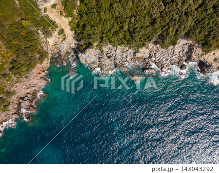Rocky bay shoreline fills frame with clear turquoise sea against forested cliffs in aquarium bay albania. Sunlit surf breaks on stones, creating vivid coastal landscape and calm, remote mood. Rocky bay shoreline fills frame with clear turquoise sea against forested cliffs in aquarium bay albania. Sunlit surf breaks on stones, creating vivid coastal landscape and calm, remote mood. 143043292