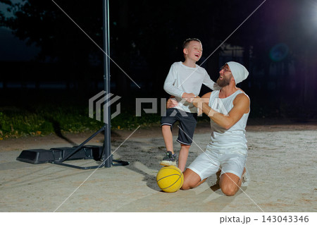 Happy family dad and son looking at camera and smiling while sitting on basketball court outdoor. Boy with handsome father after game on playground, sport concept Happy family dad and son looking at camera and smiling while sitting on basketball court outdoor. Boy with handsome father after game on playground, sport concept 143043346