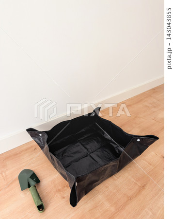 Black foldable repotting mat and green garden trowel on a wooden floor with white wall copy space 143043855
