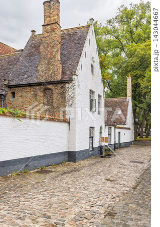 Bruges town in Belgium, Europe. Historic European street with whitewashed houses, brick walls, and cobblestone path under a cloudy sky, showcasing traditional architecture 143044667