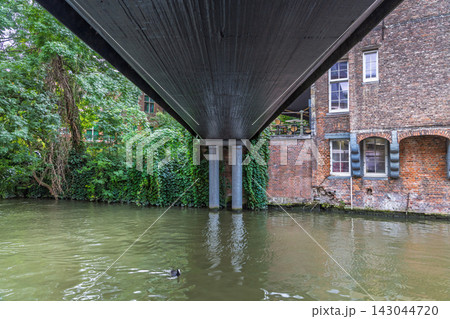 Ghent town in Belgium, Europe. View from under a bridge of a historic brick building alongside a lush, green canal, with clear water reflecting the surroundings Ghent town in Belgium, Europe. View from under a bridge of a historic brick building alongside a lush, green canal, with clear water reflecting the surroundings 143044720