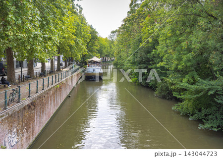Ghent town in Belgium, Europe. A serene canal scene featuring a boat docked alongside a tree-lined walkway with lush green foliage under an overcast sky Ghent town in Belgium, Europe. A serene canal scene featuring a boat docked alongside a tree-lined walkway with lush green foliage under an overcast sky 143044723