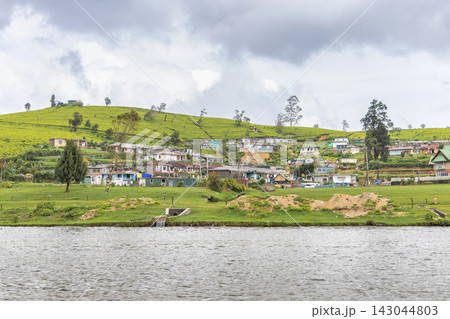Vibrant Hillside Village Overlooking Serene Lake Under Cloudy Sky - Lush Green Terraces and Rural Dwellings Reflecting Calm Waters. Nuwara Eliya town in Sri Lanka. Vibrant Hillside Village Overlooking Serene Lake Under Cloudy Sky - Lush Green Terraces and Rural Dwellings Reflecting Calm Waters. Nuwara Eliya town in Sri Lanka. 143044803