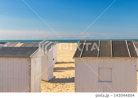 White beach huts with dark roofs line a golden sandy shore under a bright blue sky, facing a calm, distant sea expanse. Ostend, a coastal city in Belgium, Europe. White beach huts with dark roofs line a golden sandy shore under a bright blue sky, facing a calm, distant sea expanse. Ostend, a coastal city in Belgium, Europe. 143044804