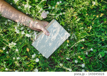 Hand holding book while relaxing on green grass with wild flowers under dappled sunlight 143045028