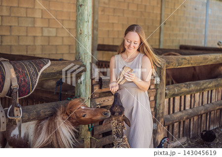 Smiling woman prepares animal feed for a pony and sheep. Joy and connection during a farm animal encounter. 143045619