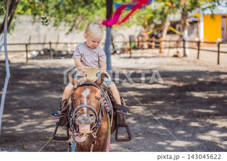 Toddler explores pony riding during a visit to a children's farm. Exploring horseback riding through a farm experience. 143045622