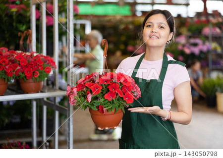 Middle-aged saleswoman holding petunia in flower-pot in open-air plants market 143050798