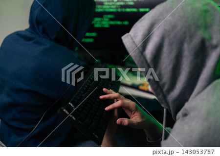 Two anonymous hackers in hoodies working together on computer. Close-up on hand typing on keyboard with green code on screen in background. Concept of cybercrime, data breach and internet security. Two anonymous hackers in hoodies working together on computer. Close-up on hand typing on keyboard with green code on screen in background. Concept of cybercrime, data breach and internet security. 143053787
