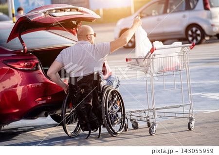 Adult disabled man in a wheelchair puts purchases in the trunk of a car in a supermarket parking lot 143055959