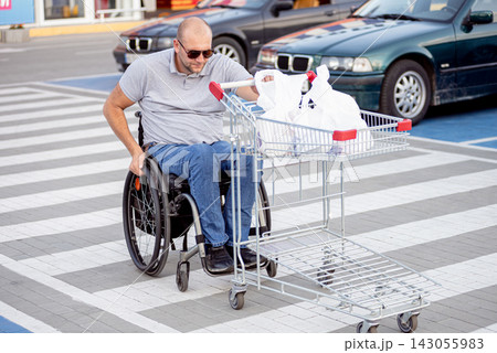 Disabled man in wheelchair pushing cart in front of himself at supermarket parking 143055983