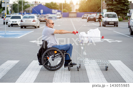 Disabled man in wheelchair pushing cart in front of himself at supermarket parking 143055985
