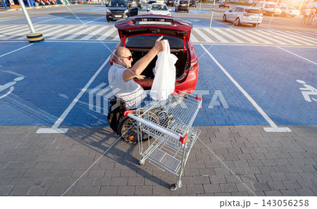 Adult disabled man in a wheelchair puts purchases in the trunk of a car in a supermarket parking lot 143056258