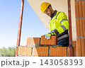 Skilled builder wearing safety helmet positioning red brick block on mortar 143058393