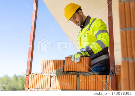 Skilled builder wearing safety helmet positioning red brick block on mortar 143058393