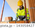 Male mason wearing helmet applying cement mixture with trowel 143058427