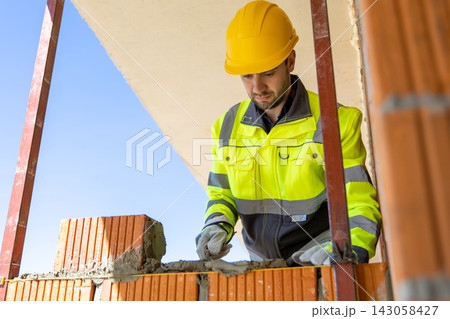 Male mason wearing helmet applying cement mixture with trowel 143058427
