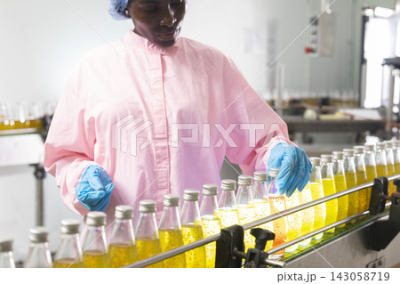 Black woman inspecting drink quality with rows of glass bottles on the automated conveyor system in manufacturing assembly line. 143058719