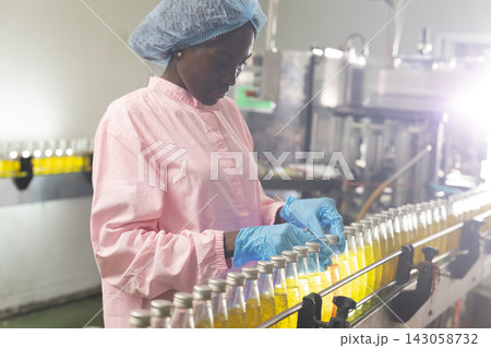 Black woman inspecting drink quality with rows of glass bottles on the automated conveyor system in manufacturing assembly line. Black woman inspecting drink quality with rows of glass bottles on the automated conveyor system in manufacturing assembly line. 143058732