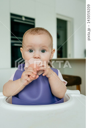 Cute baby sitting in a high chair at home and enjoying a snack. Natural indoor family moment showing early childhood, feeding, curiosity and everyday life 143059209