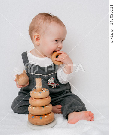 Adorable baby girl wearing gray overalls sitting on white blanket and playing with eco friendly wooden pyramid toy. Early motor skill development, eco toys, and teething period concept 143059211