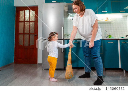 Young Caucasian educator teaches a little girl to sweep the floor with a broom in a kindergarten classroom. The Montessori activity develops practical skills, coordination, and independence. Young Caucasian educator teaches a little girl to sweep the floor with a broom in a kindergarten classroom. The Montessori activity develops practical skills, coordination, and independence. 143060581