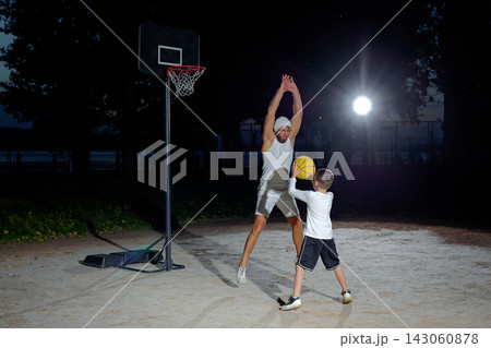 A father and son enjoy a night basketball game at a local park under bright lights focusing on teamwork and fun 143060878