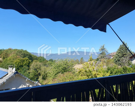 A peaceful balcony view overlooking leafy countryside, rooftops and distant Pelat massif in the French Alp mountains under a bright blue sky. Serene, sunny rural landscape relaxation and travel. 143062193