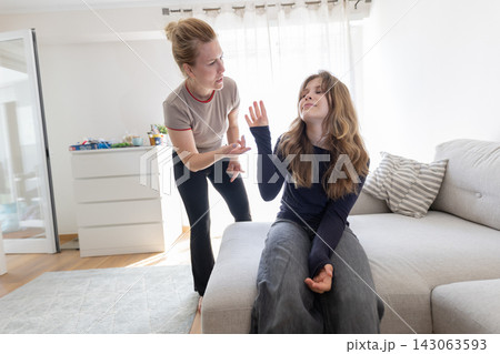 Caucasian woman talking to teenage girl sitting on a gray sofa in a bright living room, with a light-colored rug and furniture visible in the background 143063593