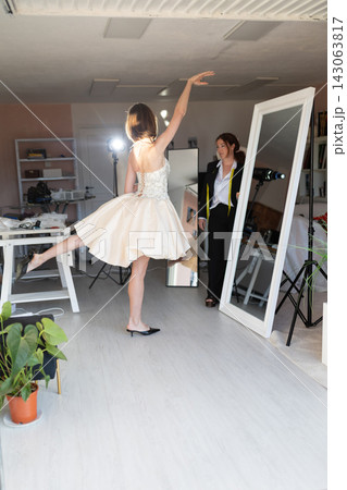 Female model wearing a short white dress poses gracefully in a photography studio with mirrors and lighting equipment visible in the background Female model wearing a short white dress poses gracefully in a photography studio with mirrors and lighting equipment visible in the background 143063817
