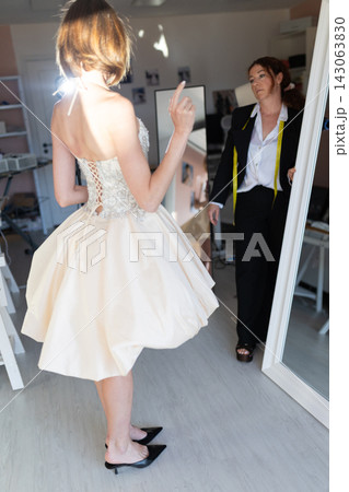 Woman in elegant cream dress poses in front of mirror while another woman in black suit observes, bright interior with mirrors and fashion elements visible Woman in elegant cream dress poses in front of mirror while another woman in black suit observes, bright interior with mirrors and fashion elements visible 143063830
