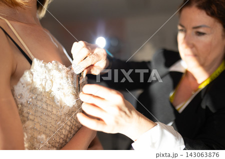 Female fashion designer adjusts intricate wedding dress on model, showcasing detailed beadwork and lace in a well-lit fitting room environment Female fashion designer adjusts intricate wedding dress on model, showcasing detailed beadwork and lace in a well-lit fitting room environment 143063876