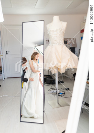 Female fashion designer adjusts wedding dress on model in front of full-length mirror, elegant bridal gown displayed on mannequin in bright studio setting 143063994