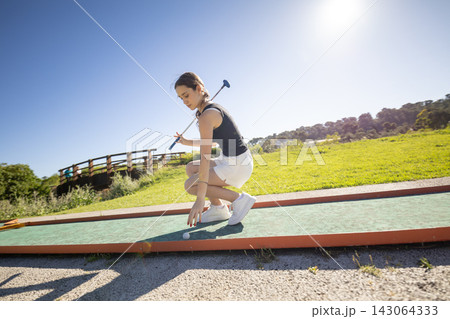 Young woman practices putting on a golf green in a park under a clear blue sky during the day 143064333