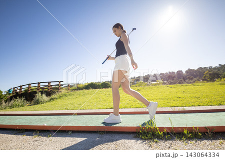 Young woman enjoys a sunny day while walking on a path with a golf club in hand near a grassy area and a wooden bridge 143064334