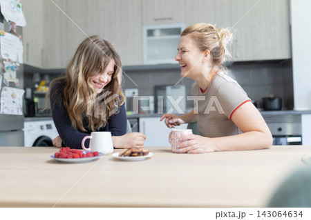 Two women laughing together at a kitchen table with coffee mugs, plates of cookies, and raspberries, in a modern home setting with appliances visible Two women laughing together at a kitchen table with coffee mugs, plates of cookies, and raspberries, in a modern home setting with appliances visible 143064634