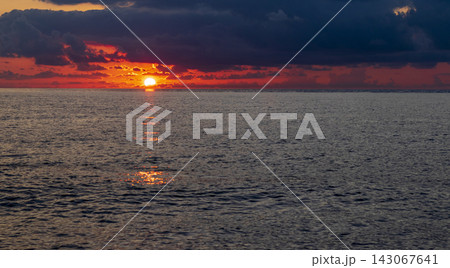 Dramatic golden sunset over the calm sea horizon with the sun bursting below dark heavy clouds. 143067641