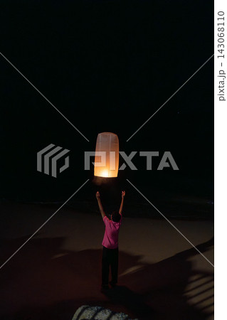 Thailand, Khao Lak, December 31, 2025: Young man releasing glowing paper sky lantern at night during festival, warm flame inside lantern lighting his hands, night sky background with soft bokeh lights 143068110