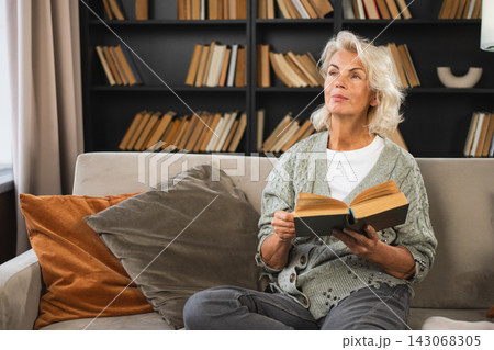Comfort relaxed senior old elderly woman reading book at home. Mature woman resting enjoy calm day sitting on sofa at home alone. Adult lady spending free time keeping book smiling. Pension concept Comfort relaxed senior old elderly woman reading book at home. Mature woman resting enjoy calm day sitting on sofa at home alone. Adult lady spending free time keeping book smiling. Pension concept 143068305