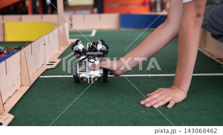 Child hand steadies compact wheeled robot on green indoor track beside wooden barriers, showing hands on robotics learning, coding practice, STEM education, and teamwork in classroom challenge area Child hand steadies compact wheeled robot on green indoor track beside wooden barriers, showing hands on robotics learning, coding practice, STEM education, and teamwork in classroom challenge area 143068462