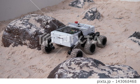 Small robotic rover with six wheels moves across sandy rock terrain beside weathered boulders, highlighting exploration technology, mobility testing, engineering design, and rugged outdoor research Small robotic rover with six wheels moves across sandy rock terrain beside weathered boulders, highlighting exploration technology, mobility testing, engineering design, and rugged outdoor research 143068468