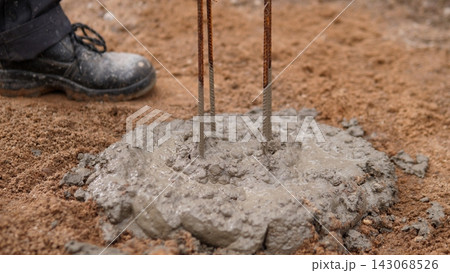 Close up view of wet concrete spreading around rusted steel rebar on sandy soil, with muddy work boot nearby, capturing foundation work, construction progress, rough texture, and hands on building Close up view of wet concrete spreading around rusted steel rebar on sandy soil, with muddy work boot nearby, capturing foundation work, construction progress, rough texture, and hands on building 143068526